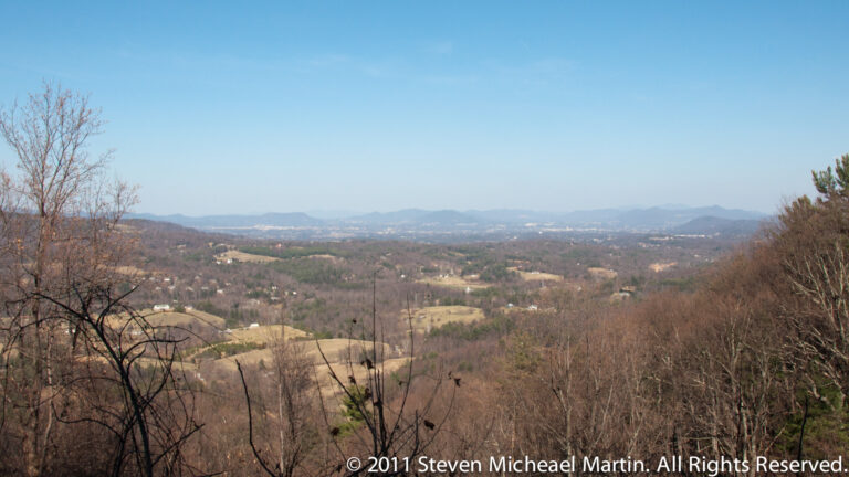 Leafless trees frame a wide view over farm fields and wooded ridges toward hazy mountains beneath a clear blue sky.