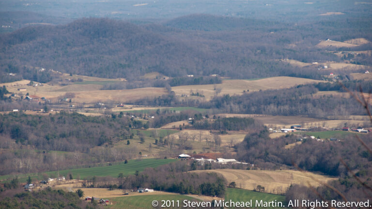 An elevated view looks over tan and green farm fields, scattered buildings, and wooded hills in a broad valley.