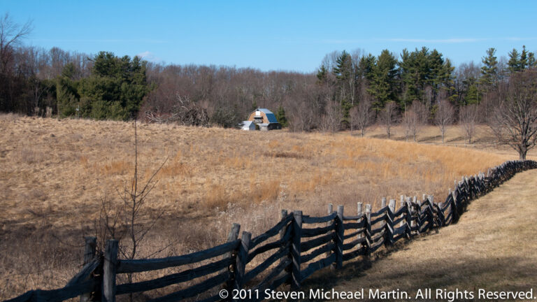 A dark zigzag rail fence crosses a tawny meadow toward a small roofed outbuilding and trees.