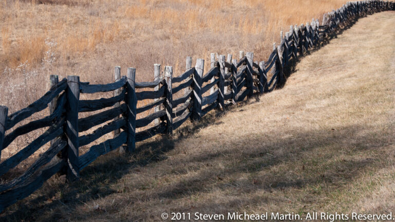 A weathered zigzag fence of dark rails and gray posts recedes across a dry grassy field.