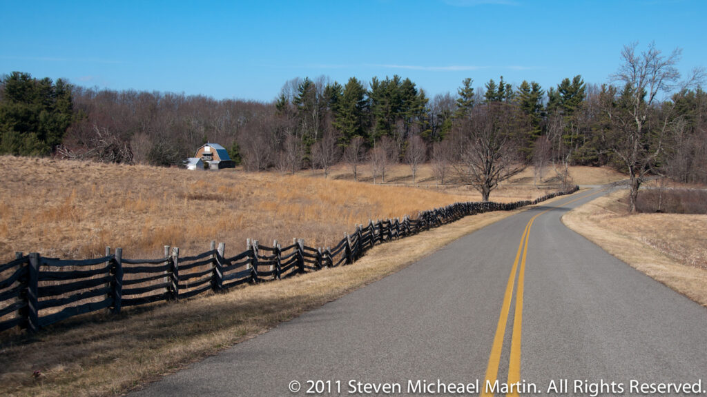 A paved road with double yellow lines bends through dry pasture beside a rail fence and scattered trees.