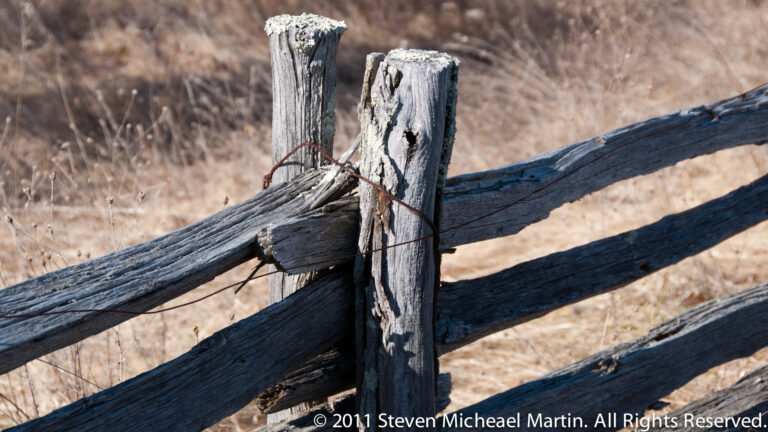 Close-up of weathered gray fence posts and rails joined with a loop of rusty wire.