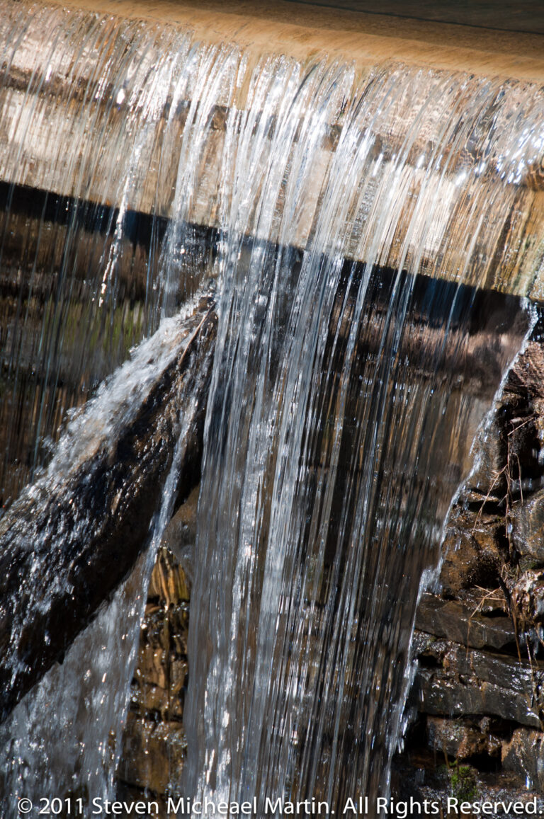 Water pours over a low curved weir in narrow shimmering streams onto dark rocks below.