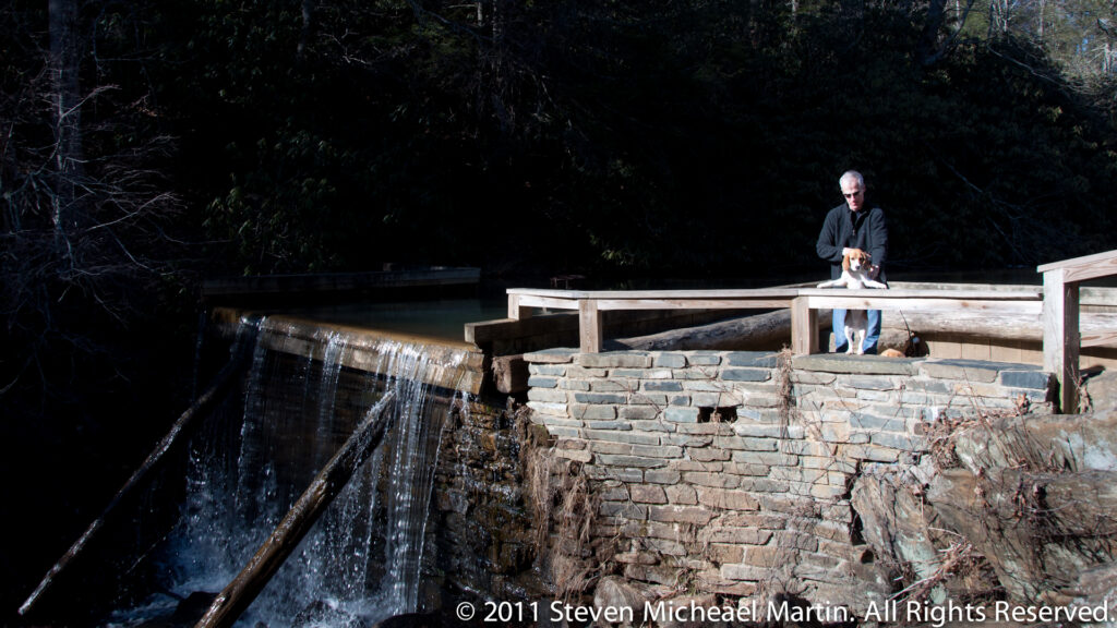 A person with a small dog stands on a wooden footbridge beside water spilling over a low stone weir.