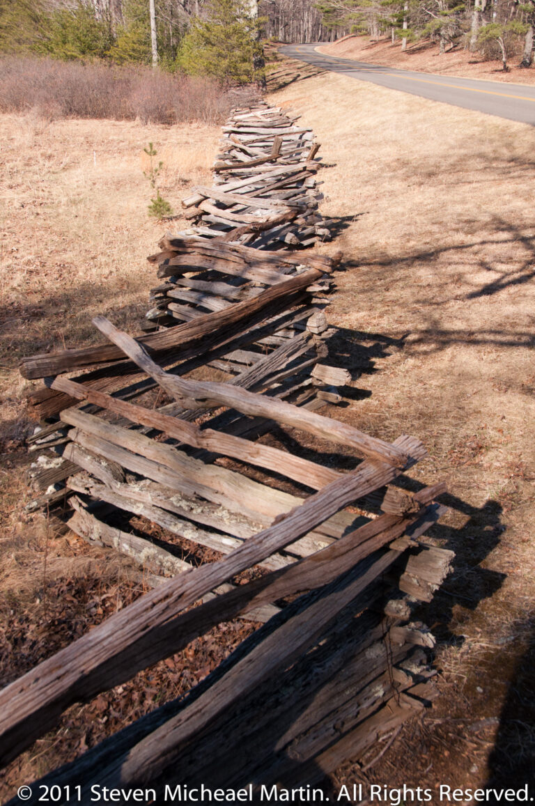 A heap of weathered wooden fence rails lies on dry ground beside a paved road.