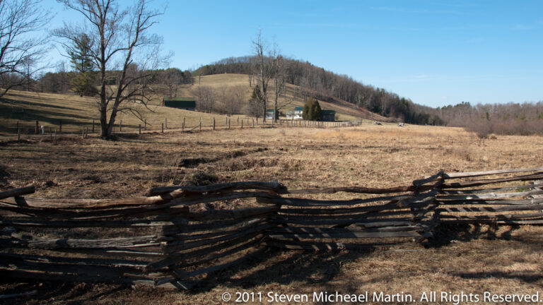 A broken rail fence crosses the foreground of an open pasture, with farm buildings and a wooded hill beyond.
