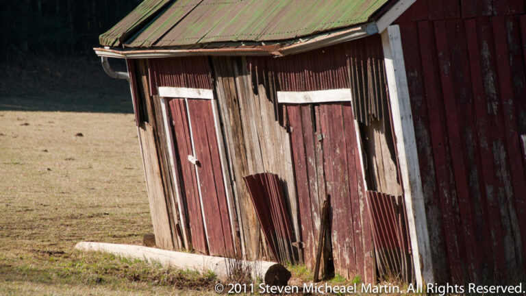 Weathered red and brown barn boards rise beneath a patchy green metal roof, with a small white-trimmed opening.