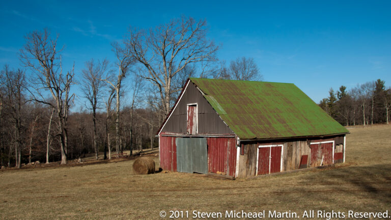 A weathered red barn with a green roof stands in a dry field beside a round hay bale.
