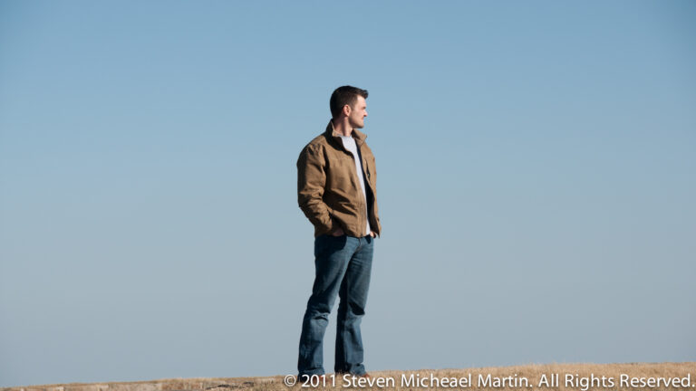 A man in a brown jacket and jeans stands on a low grassy ridge, looking to the side beneath a broad blue sky.