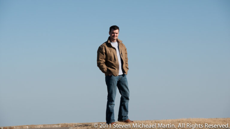 A man wearing a brown jacket and jeans stands on a narrow grassy ridge against a large blue sky.