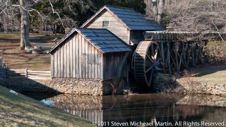 A wooden mill building with a large waterwheel stands over a narrow stream beside a smaller outbuilding.