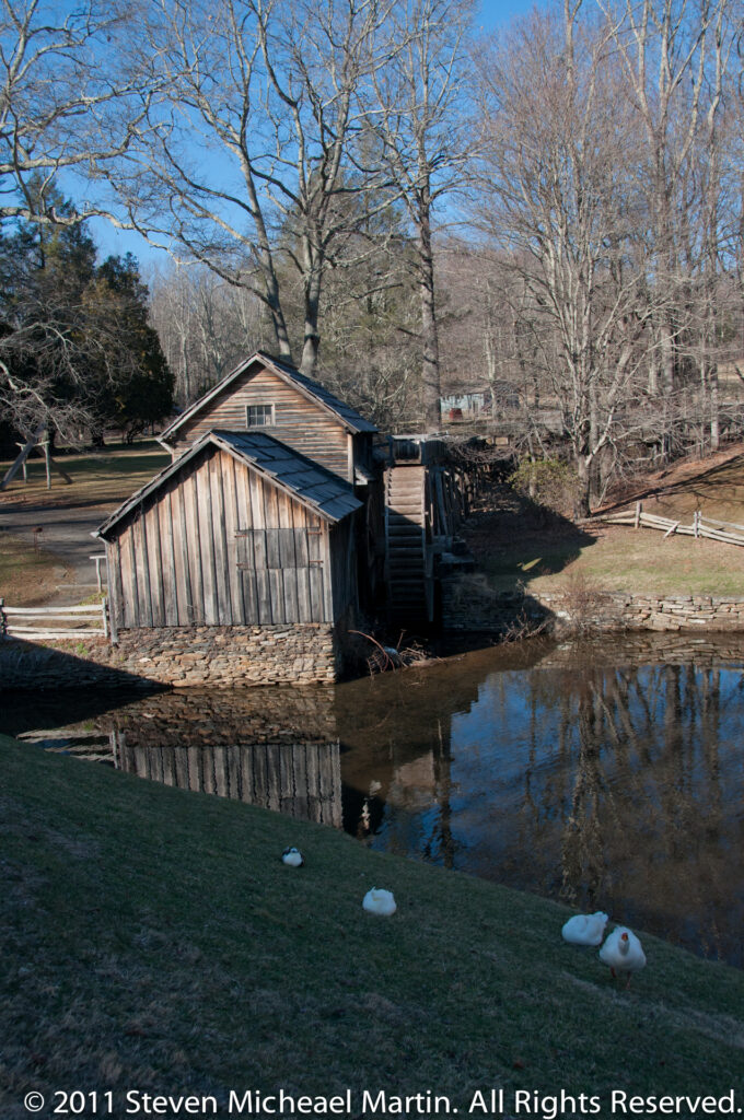 A wooden mill and large waterwheel reflect in a pond, with white waterfowl resting along the grassy shore.