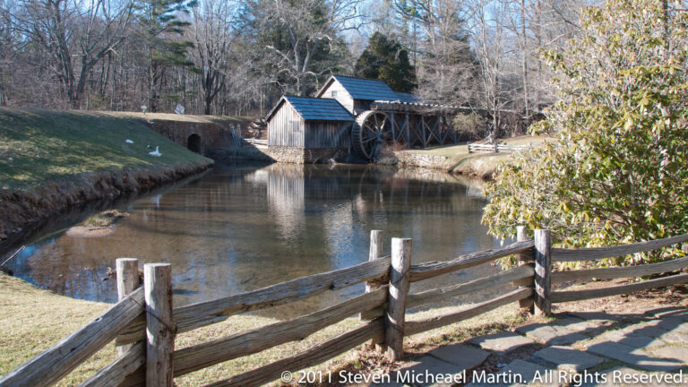 A stone path and wooden fence lead beside a pond toward a timber mill and waterwheel.