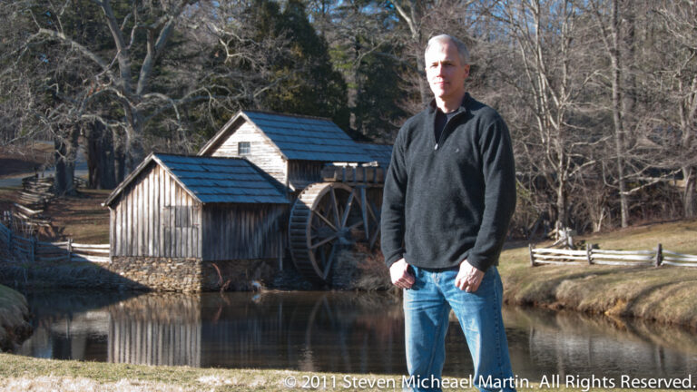 A man in a dark sweater stands beside a pond, with a wooden mill and waterwheel behind him.