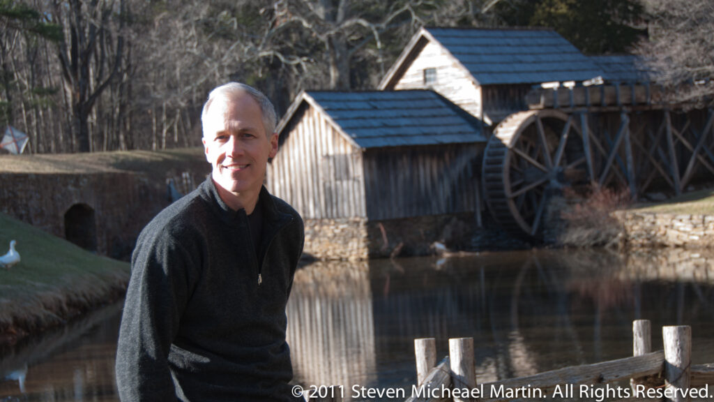 A visitor stands beside Mabry Mill’s large wooden waterwheel, with timber buildings and the pond behind him.