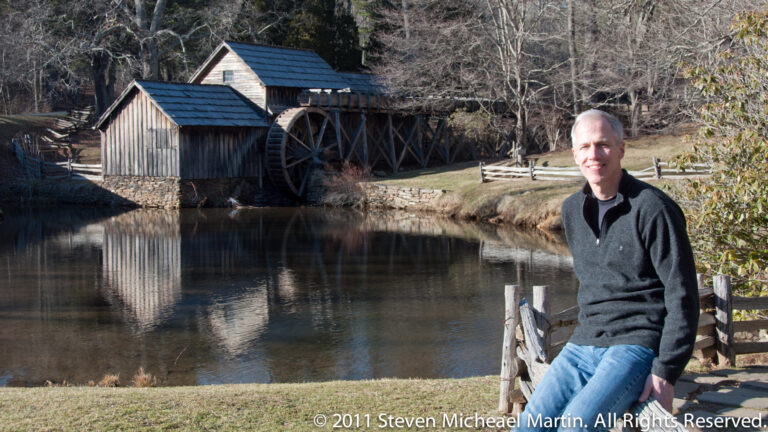A seated visitor is beside a pond reflecting Mabry Mill’s timber buildings and large waterwheel.