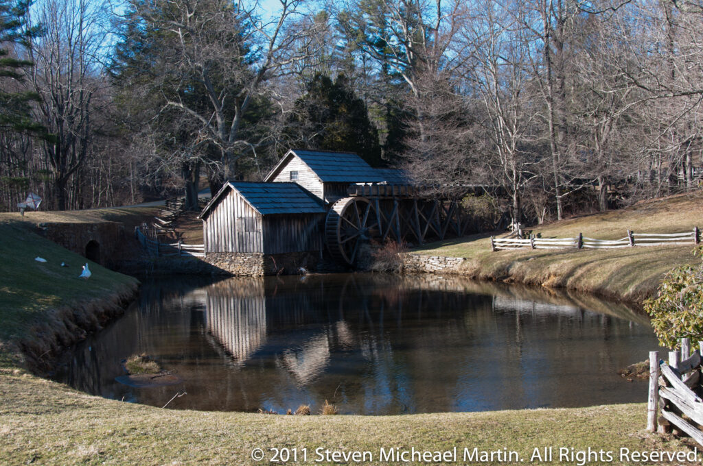 Mabry Mill’s timber buildings and waterwheel reflect in a still pond bordered by trees.