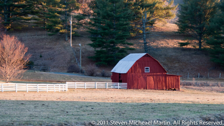 Red gambrel-roof barn stands behind a white fence in an open pasture below wooded hills.