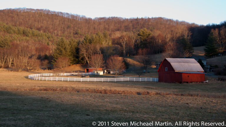 Wide pasture with a red barn, white fences, distant cattle, and a wooded ridge in warm evening light.