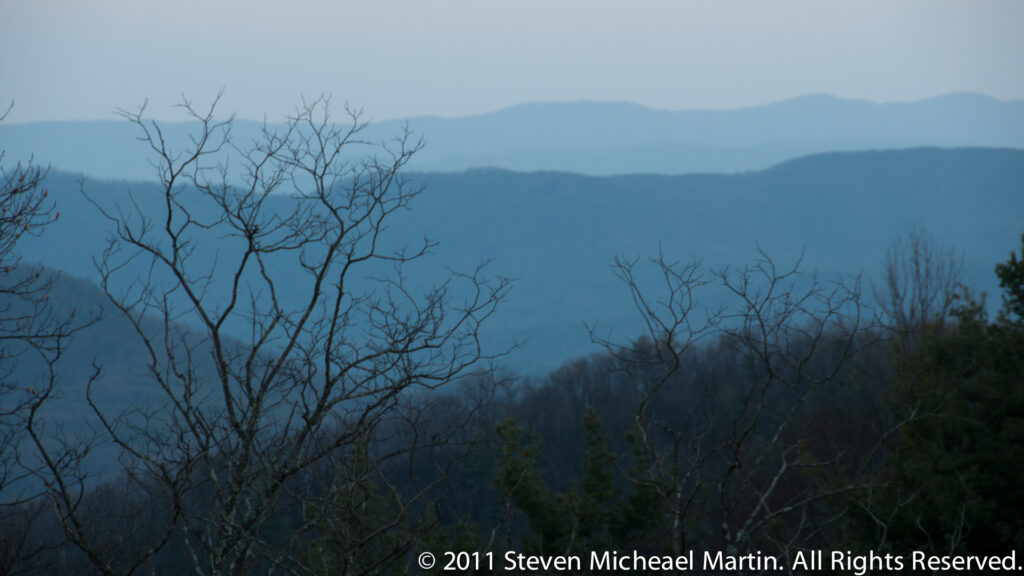 Bare branches frame hazy Blue Ridge ridgelines fading into a pale sky above dark foreground forest.