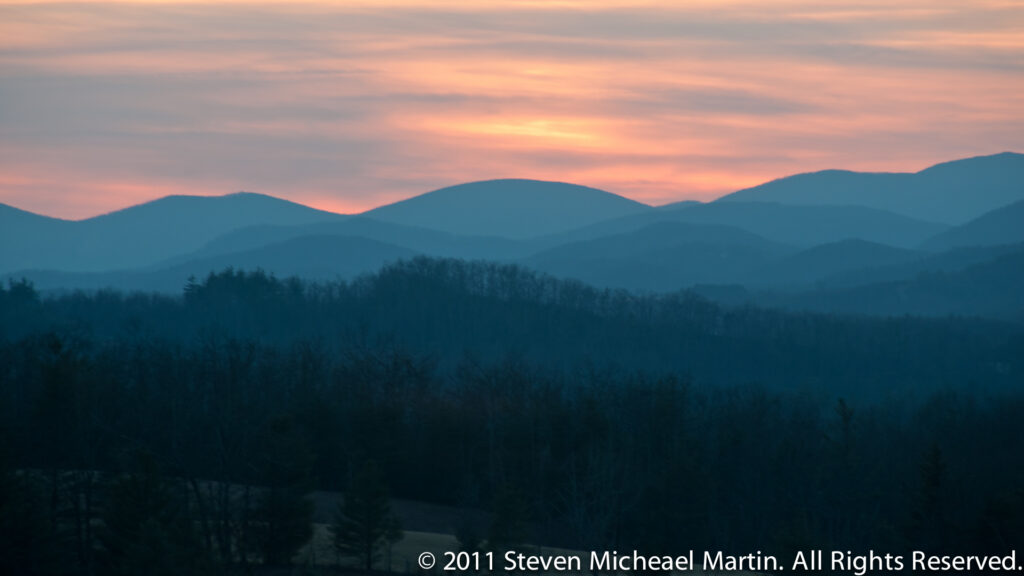 Coral sunset sky glows above layered blue ridges and a dark wooded foreground.