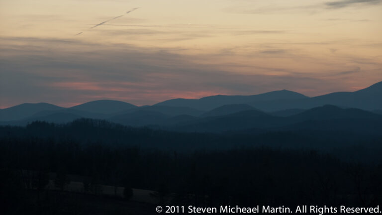 Muted peach-gray clouds hang above layered blue ridges and dark forest silhouette.