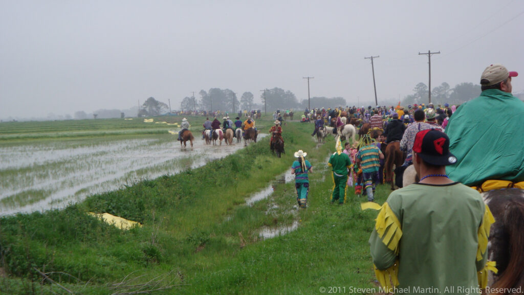 A line of costumed riders and walkers follows a grassy track between flooded fields in mist.