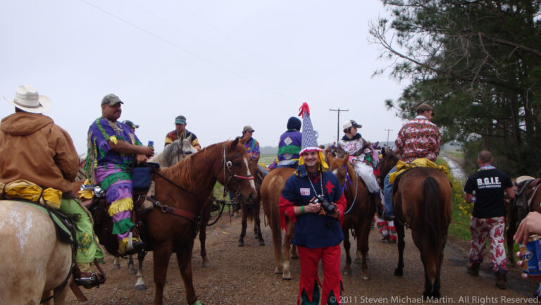 Costumed participants stand among closely grouped horses on a muddy track at a rural Mardi Gras gathering.