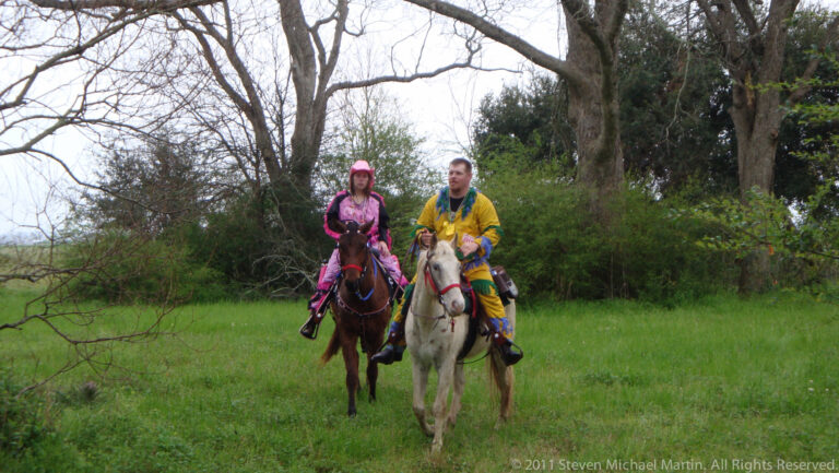 Two costumed riders approach on a brown horse and a light-colored horse across a field.