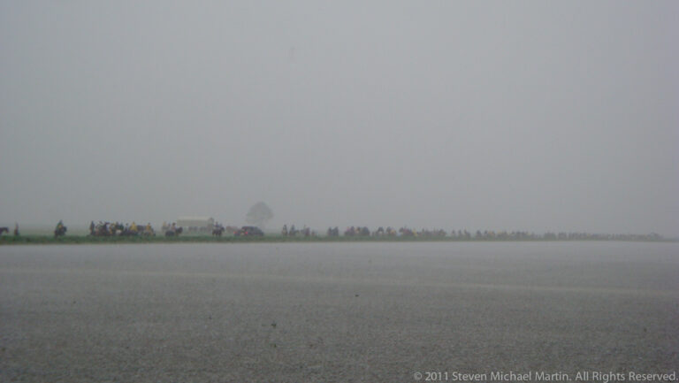 Distant riders move along a narrow grassy berm between flooded fields in dense fog.