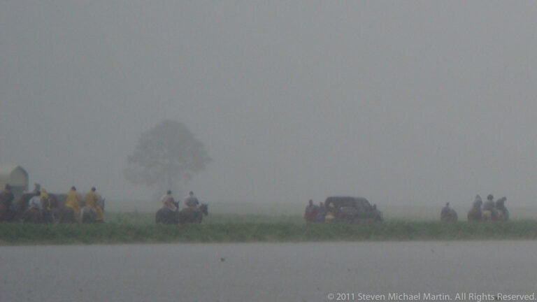 Small groups of riders and a vehicle gather on a grassy levee above flooded fields in fog.