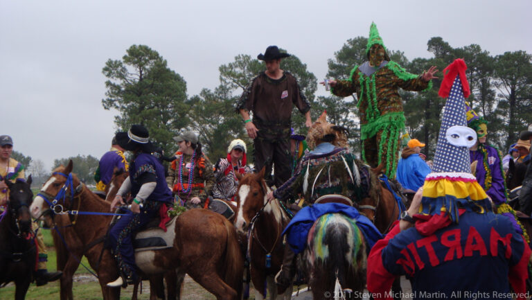 A participant in a green fringed costume stands among riders gathered on horses in a field.