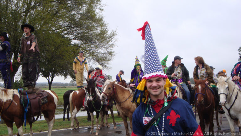 A smiling man in a tall blue-and-white pointed hat stands before a group of costumed riders.