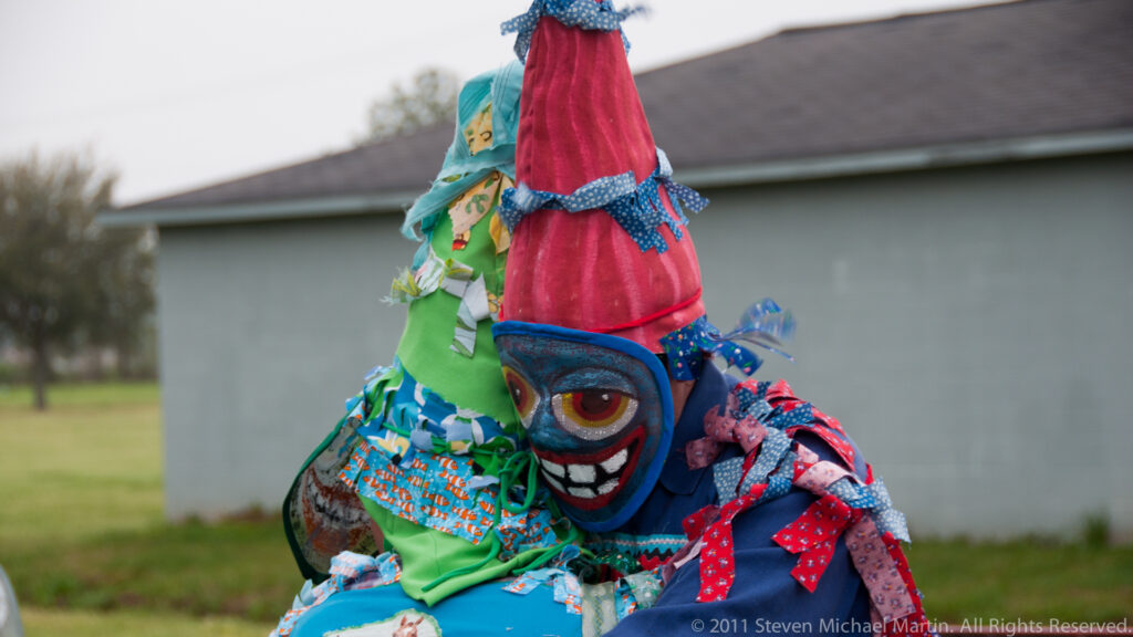 A participant wears a colorful grinning mask and layered fabric costume beside a building.