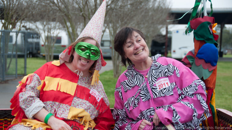 Two smiling women pose together, one in a green eye mask and cone hat and the other in pink.