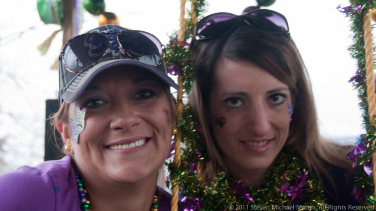 Two women smile beneath Mardi Gras decorations, wearing beads, a cap, and small face decorations.