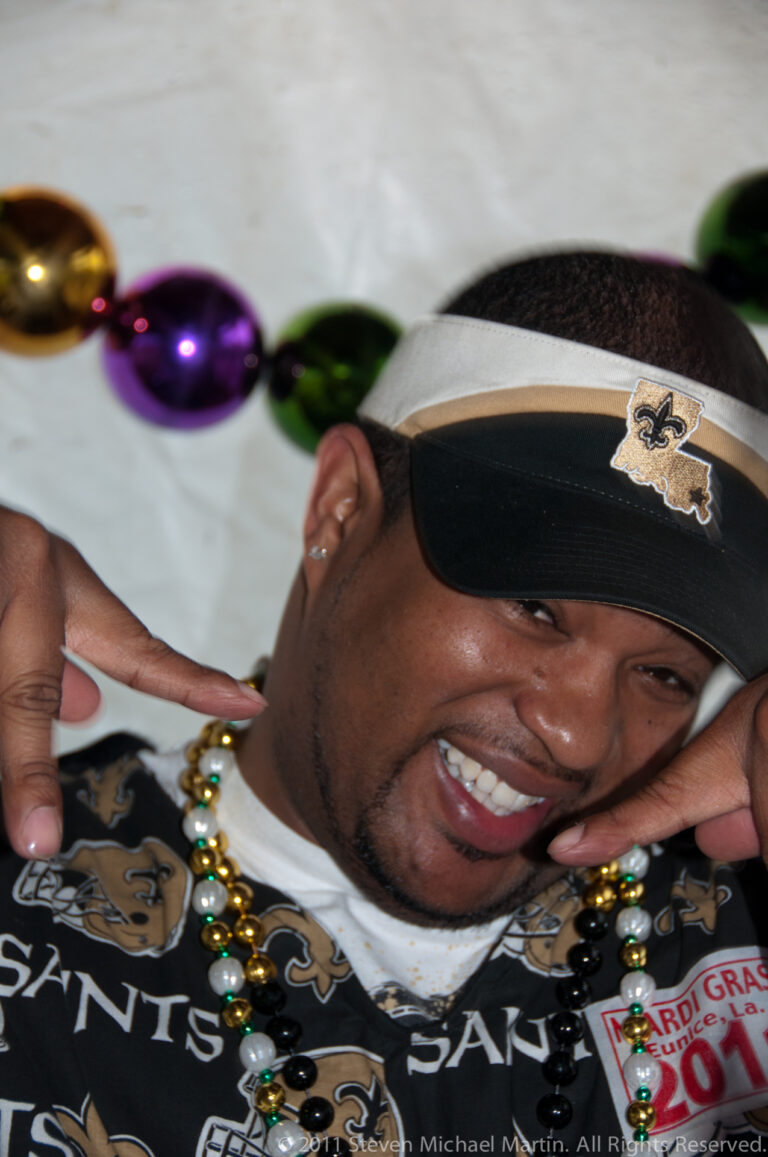 A smiling man in Saints apparel poses with beads and a Mardi Gras event badge.