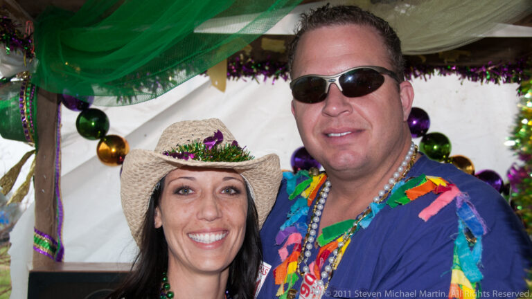 A smiling couple pose beneath a decorated canopy, wearing beads, sunglasses, and a straw hat.