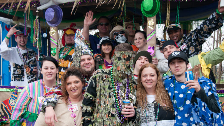 A crowded group in colorful costumes waves beneath a bead-decorated trailer canopy.