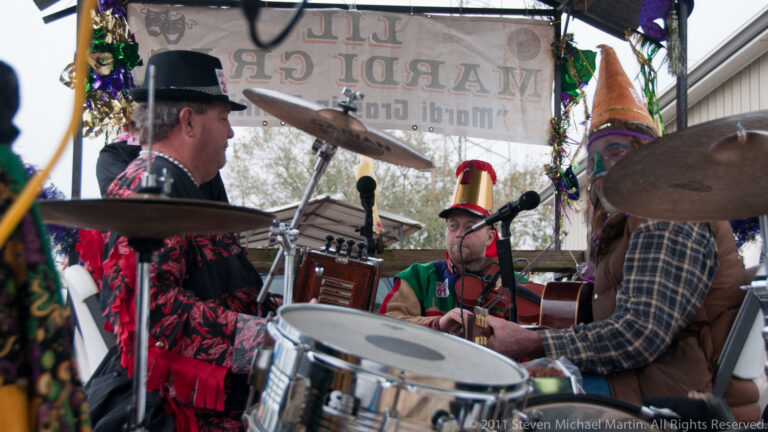 Musicians play drums and string instruments beneath a decorated canopy on a trailer.