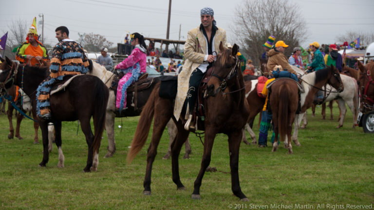 Costumed riders and horses crowd together in a field, with a man in a light jacket near the center.
