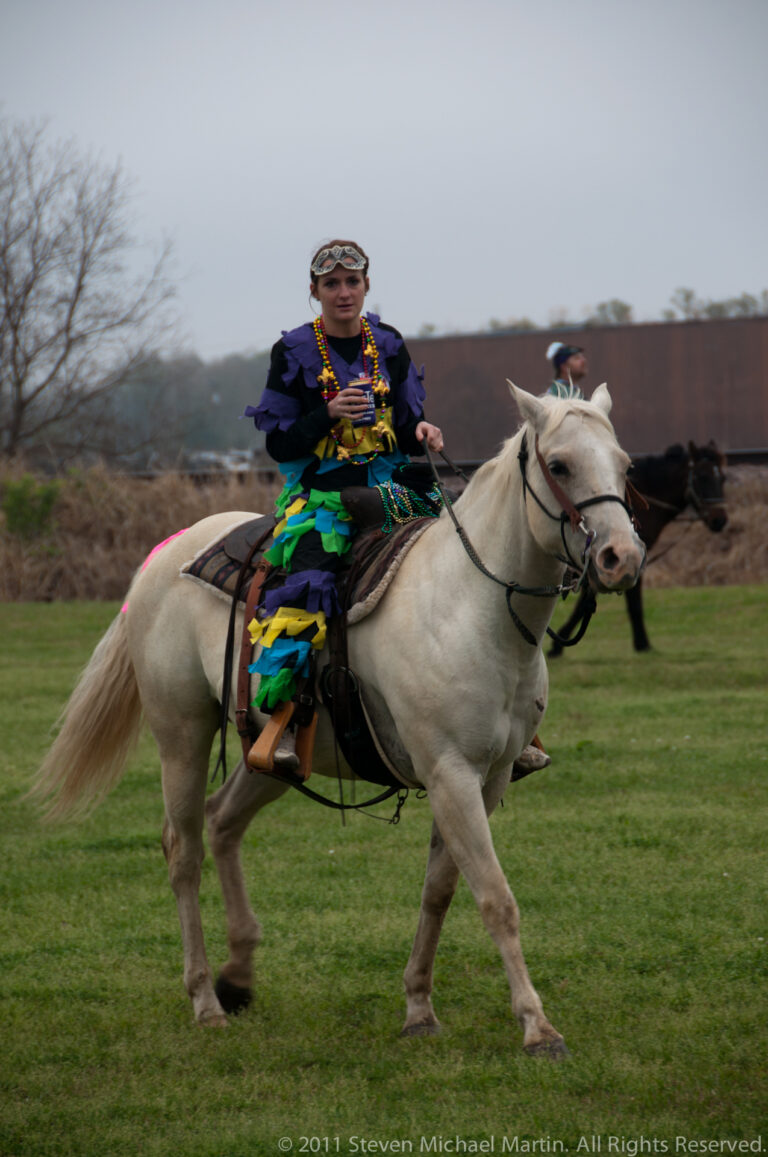 A costumed participant rides a white horse among other riders, with freight cars in the distance.
