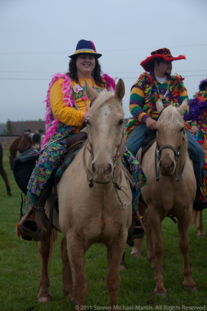 Two costumed riders approach on horses, with other mounted participants and a grassy field behind them.
