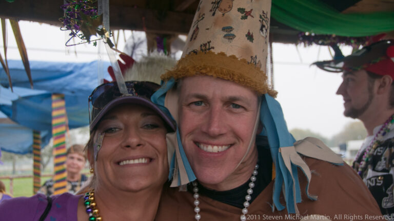 Smiling couple poses beneath a decorated shelter; the man wears a tall pointed hat and beads.