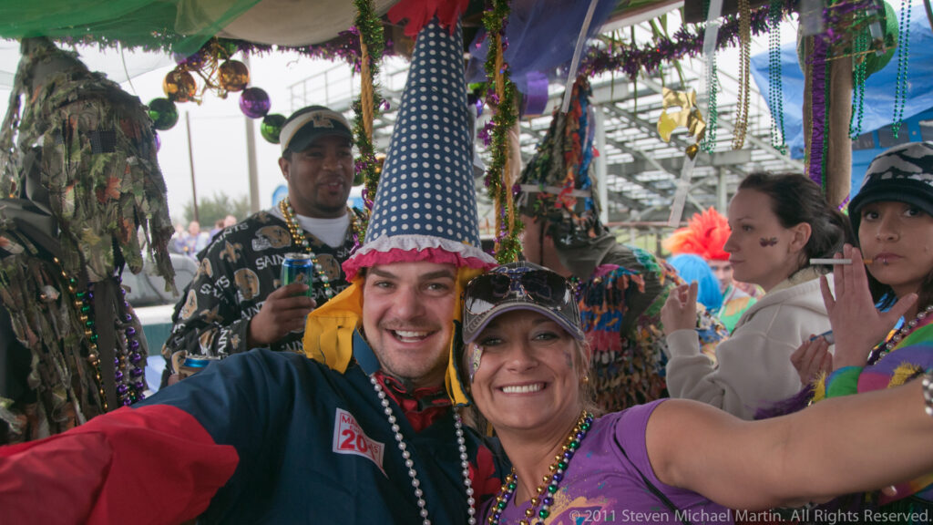 Man in a tall patterned hat and woman pose for a selfie among festivalgoers beneath a canopy.