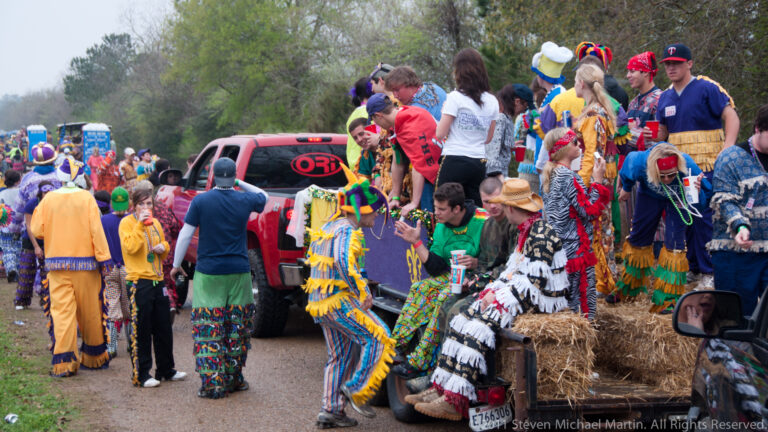 Costumed crowd surrounds a red pickup and hay-covered trailer on a wet rural road.