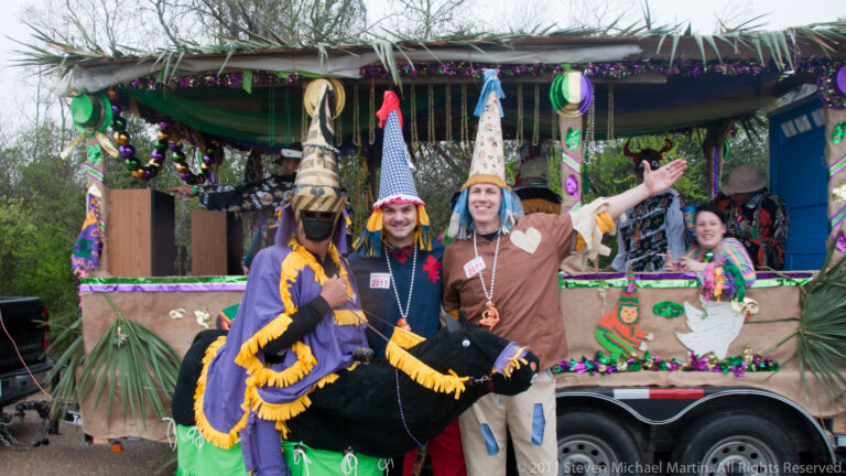 Costumed participants pose beside a trailer decorated with greenery, beads, and hanging Mardi Gras ornaments.