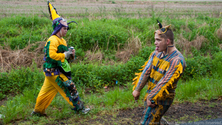 Two people in yellow and green, and striped patchwork costumes, walk along a muddy grassy path.