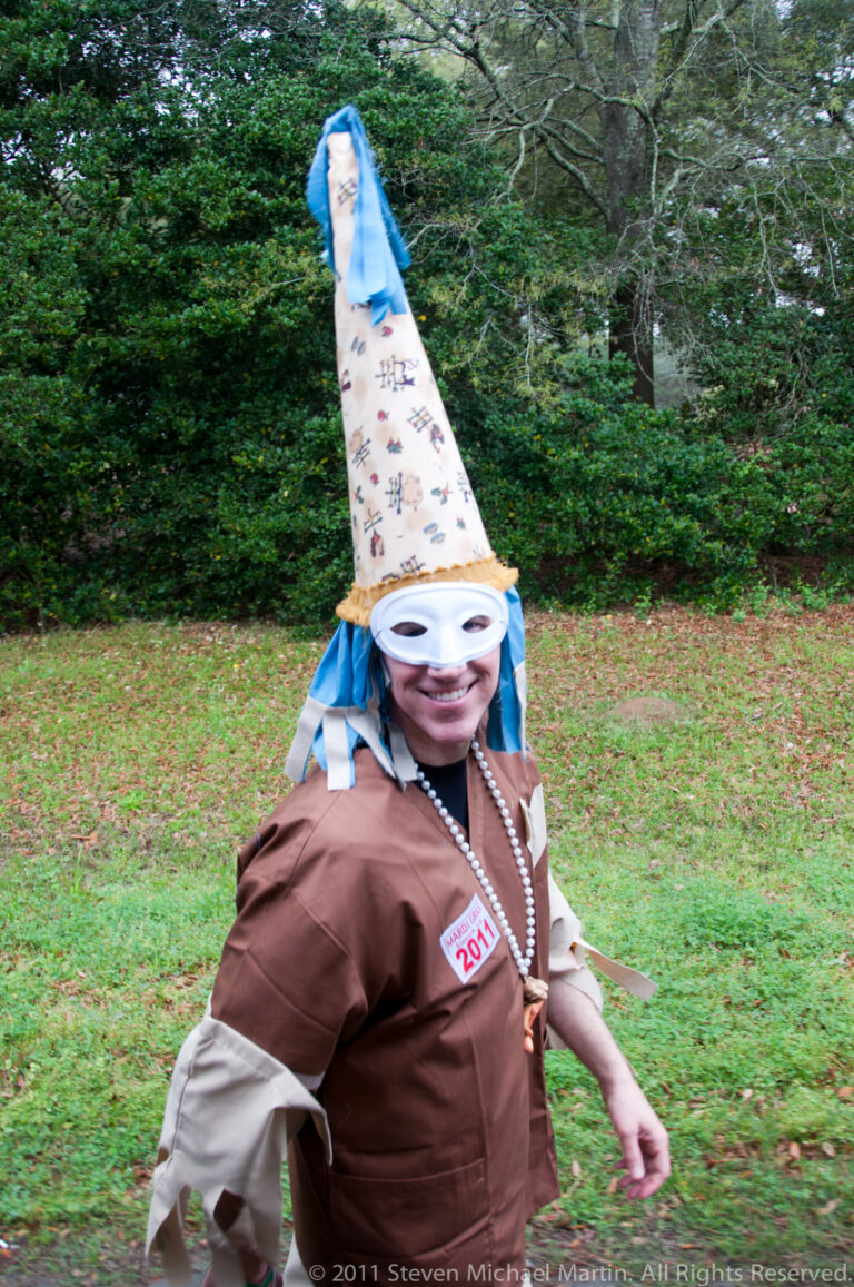 Smiling participant wears a decorated pointed hat, oversized white goggles, beads, and a brown costume.