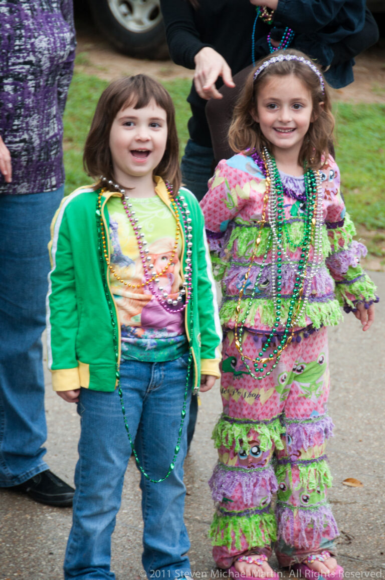Two girls in green and pink outfits stand together wearing many strands of colorful beads.
